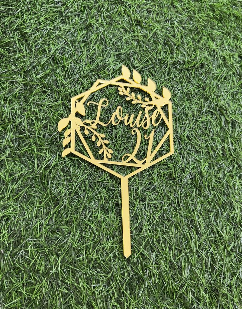 A golden acrylic cake topper displayed on a green artificial grass background. The topper features a geometric frame adorned with leaves and flowers, with the text "Louise" and the number "21" artistically integrated within the design. The topper&