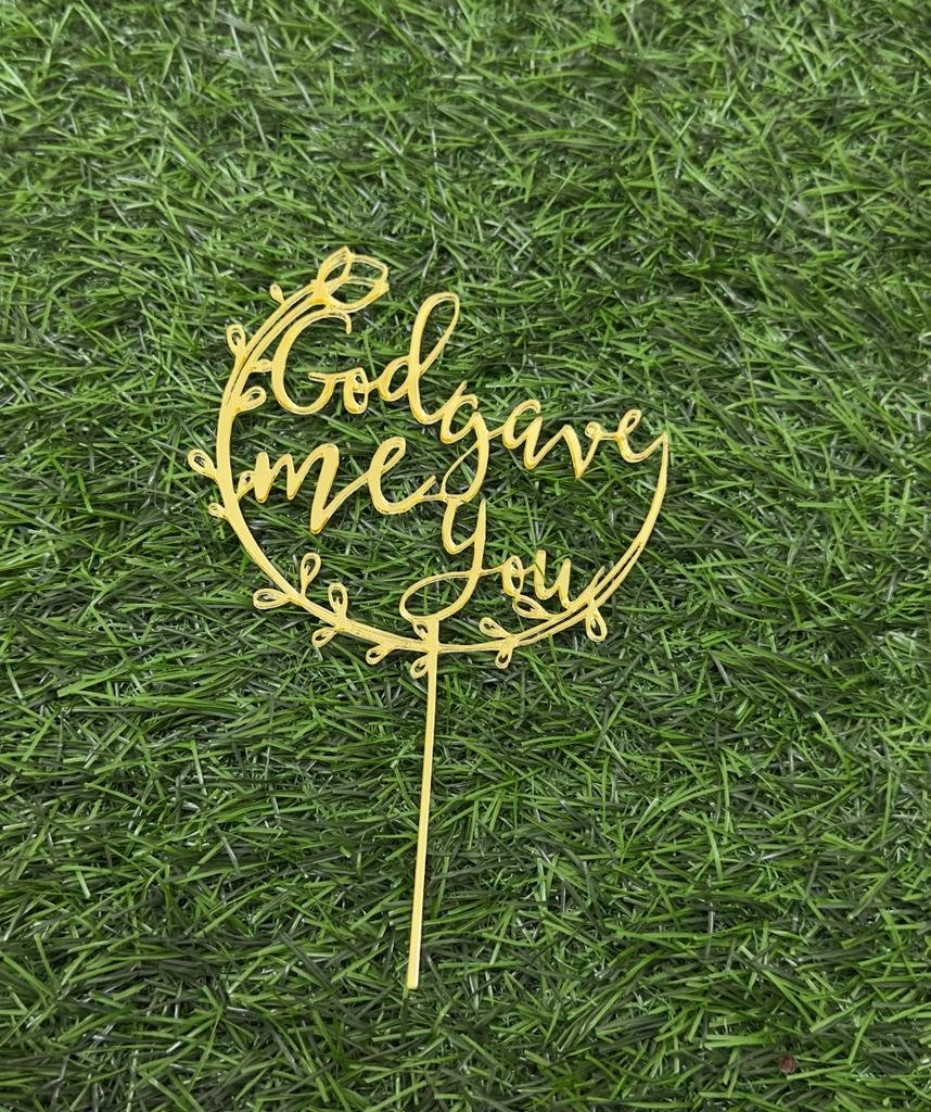 A gold acrylic cake topper displayed on a green grass-like surface. The topper features the phrase "God gave me You" in an elegant, scripted font, surrounded by decorative leaf motifs. It is designed for use in cake decorations.