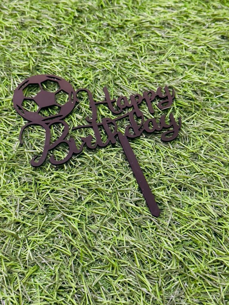 A cake topper featuring the phrase "Happy Birthday" in an elegant script. It is designed with a soccer ball at the beginning of the text. The topper is placed on a green grass-like background, emphasizing its decorative purpose for a birthday celebration.