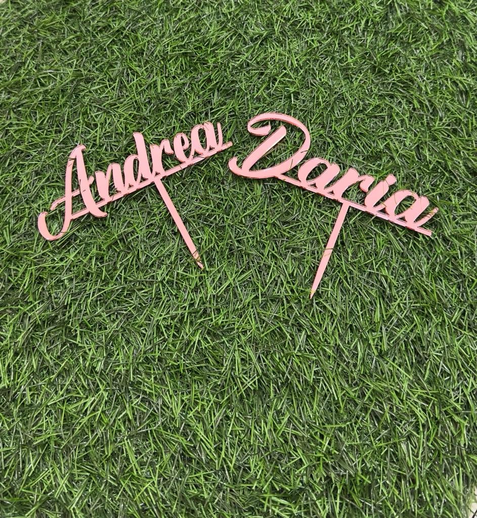 Two customized cake toppers with the names "Andrea" and "Daria" written in elegant cursive font. The toppers are made of a pink material and are placed on artificial grass.