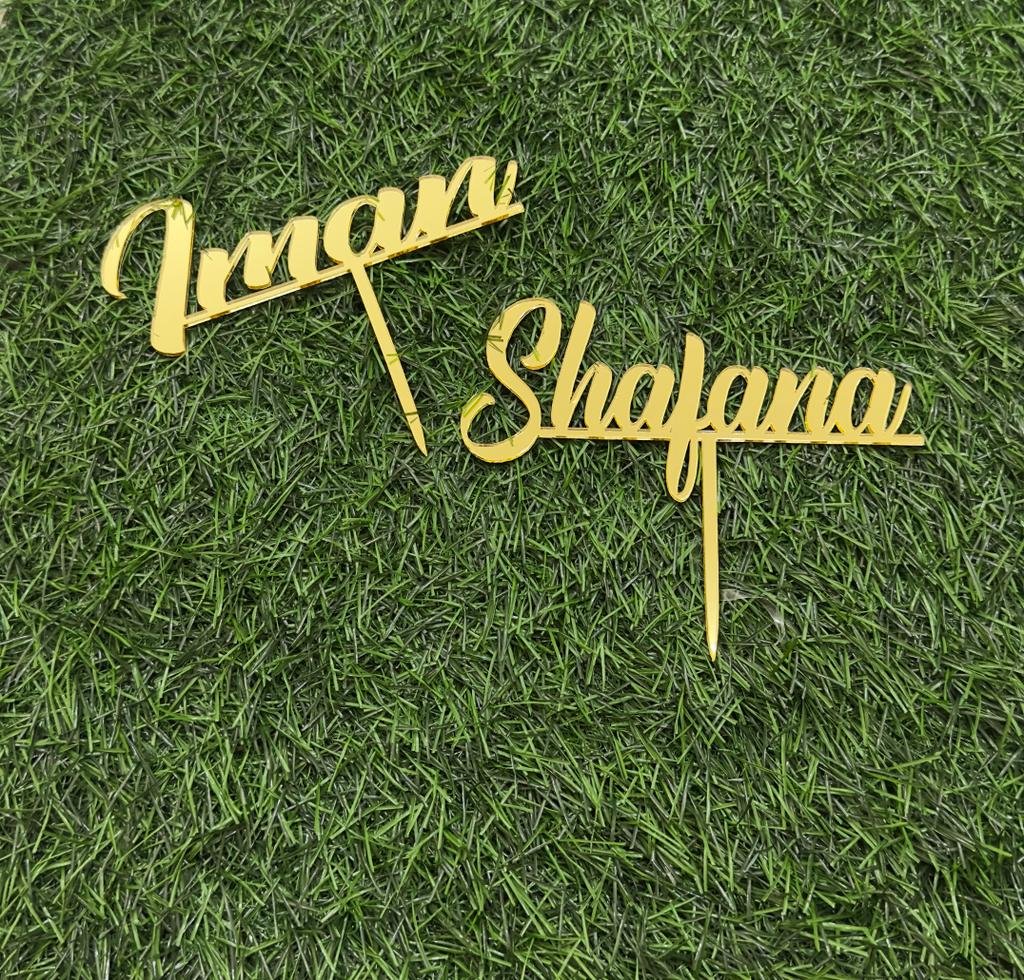 Two gold cake toppers with the names "Imran" and "Shafana" displayed on a green grass-like background. The toppers feature an elegant, cursive font with pointed ends for easy insertion into a cake.