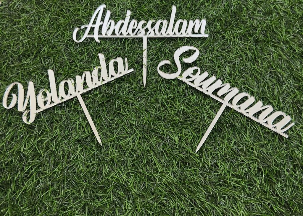 Three custom cake toppers placed on a grassy surface. The toppers are made of a light-colored material and feature the names "Abdessalam," "Yolanda," and "Sourmana" in a decorative cursive font. Each topper has a pointed end for insertion into a cake.