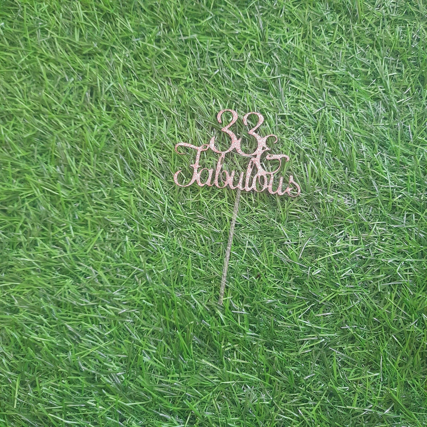 A gold glitter card stock cake topper that reads "33 Fabulous," resting on a green artificial grass background. The topper features a decorative font with a stick for placing in a cake.