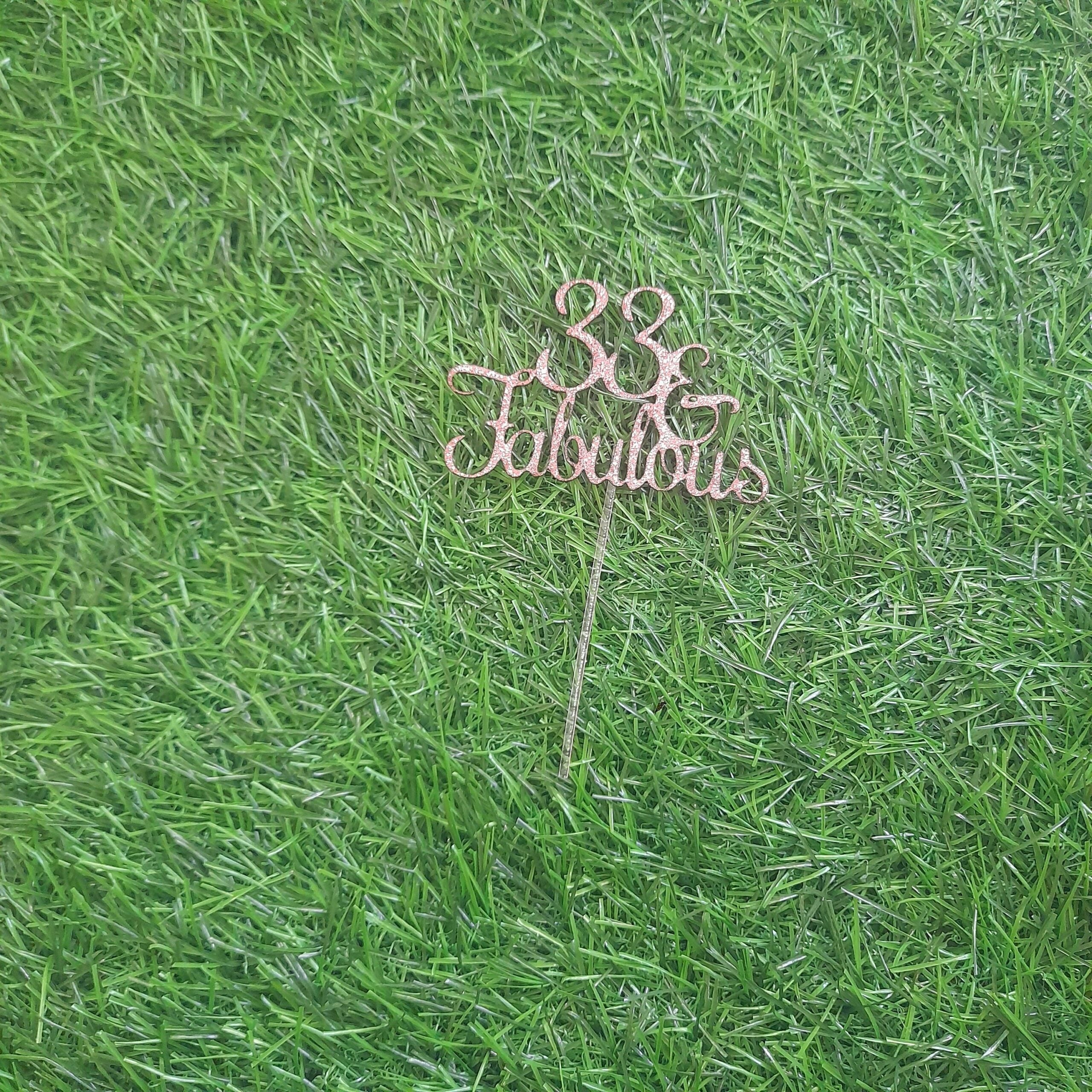 A gold glitter card stock cake topper that reads "33 Fabulous," resting on a green artificial grass background. The topper features a decorative font with a stick for placing in a cake.