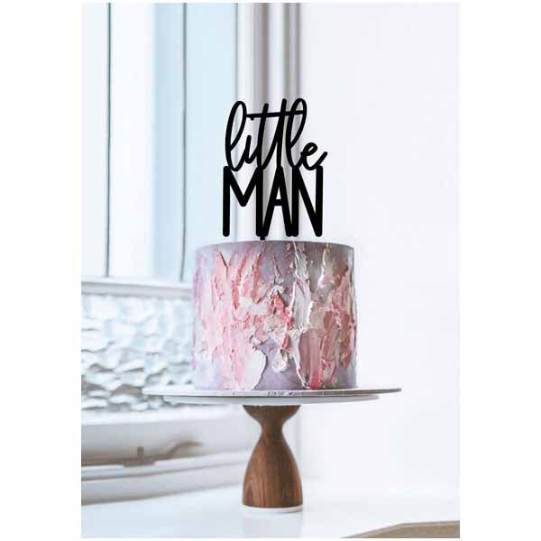 A round cake with a textured, swirled frosting that features shades of pink and white. Atop the cake is a black acrylic topper with the words "little MAN" in a playful font. The cake is displayed on a simple white cake stand with a wooden base, positioned near a window with soft natural lighting.