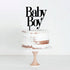 An unfinished, two-layer white cake with a rustic naked design, displaying hints of brown in the frosting, sits on a wooden cake stand. Topping the cake is a black acrylic cake topper that reads "Baby Boy" in playful typography. The background is light and neutral, enhancing the focus on the cake.
