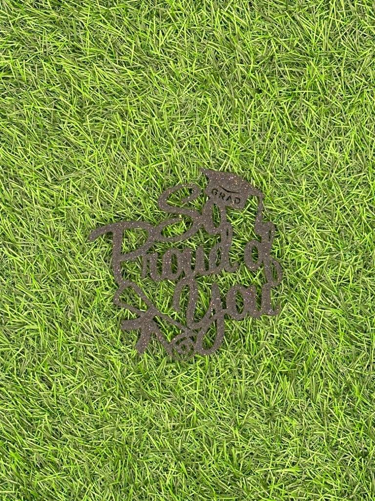 A black cardstock cake topper design placed on green grass. The topper features the words "So" and "Thank You," with a graduation cap above the word "Grad," and a decorative element resembling a flower or star.
