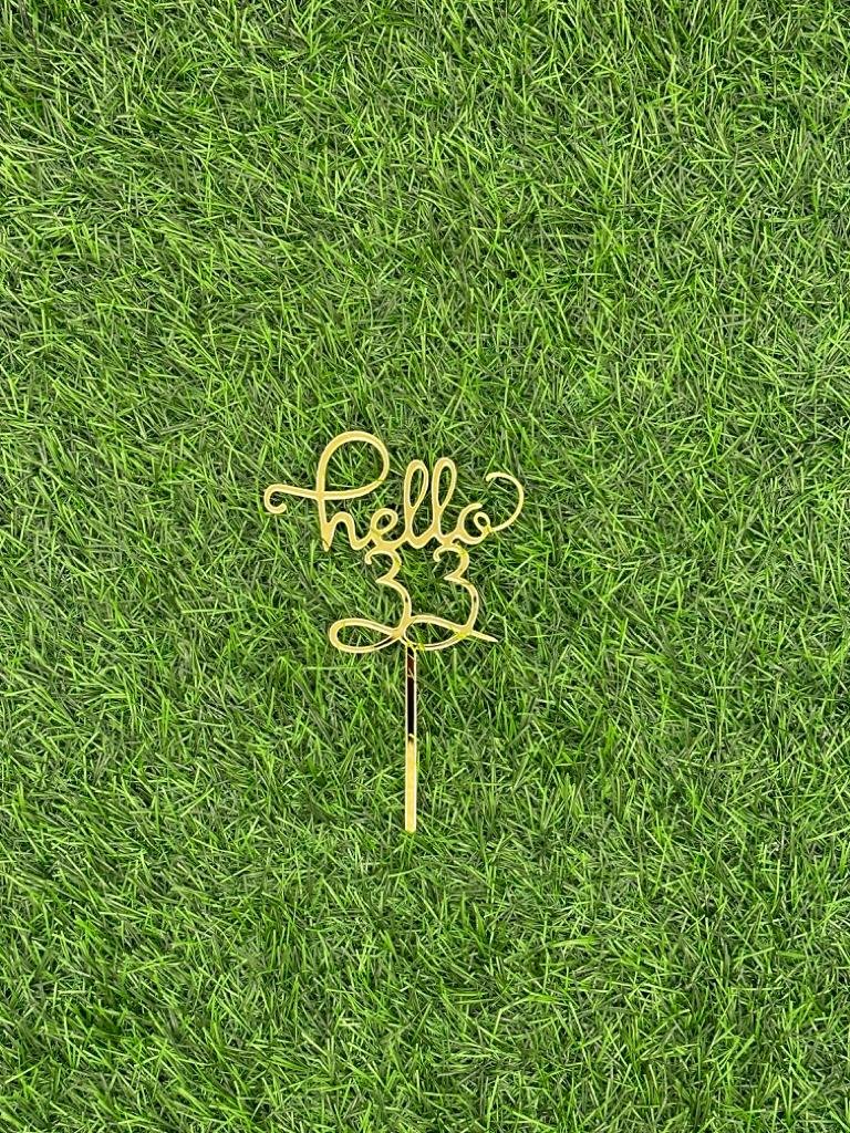 The image shows a gold-colored cake topper with the word "hello" and the number "33" styled in a decorative script. The topper is placed against a backdrop of artificial green grass. The design is intended for use in cake decoration, possibly for a celebration.