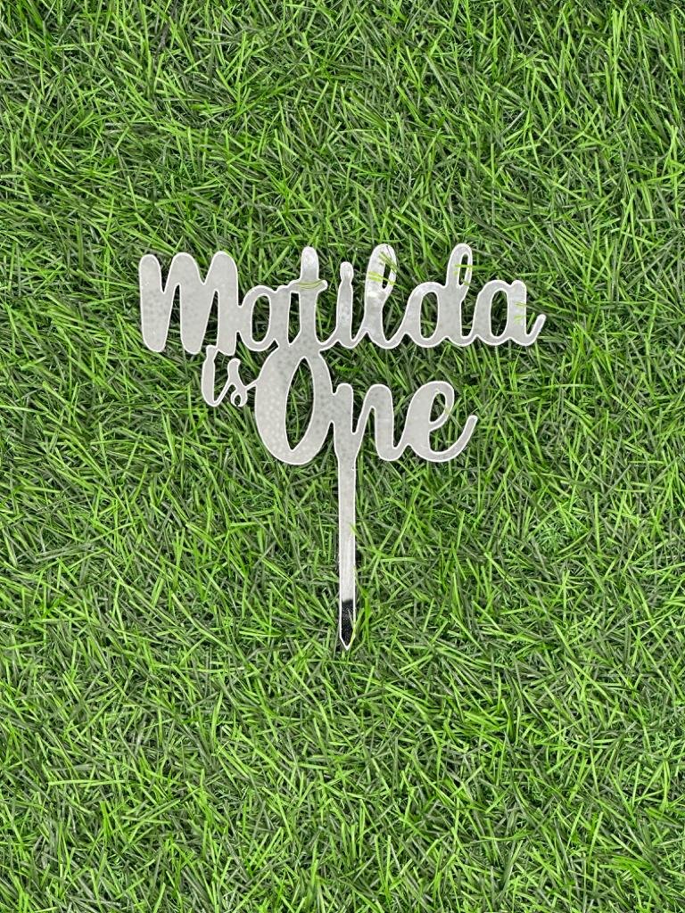 A decorative acrylic cake topper featuring the text "Matilda is One" in a stylish script font. The topper has a stake for easy insertion into a cake. It is displayed against a background that resembles green grass or artificial turf.