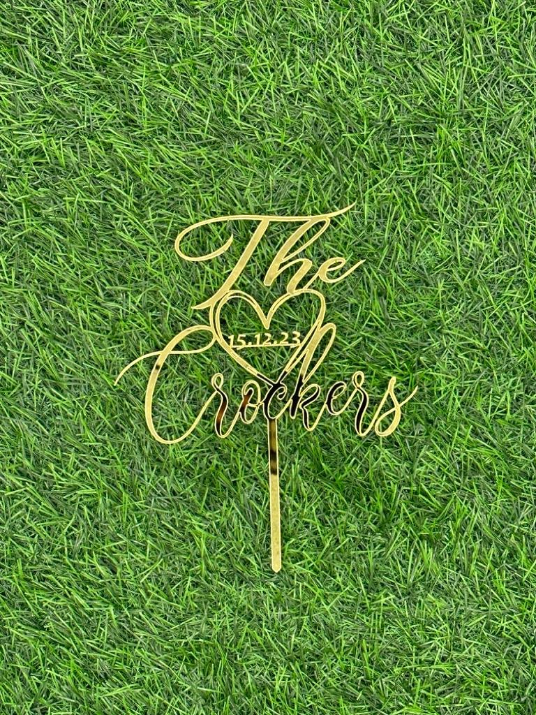 A golden acrylic wedding cake topper is displayed on a green grass background. The topper features the text "The Crockers" with a heart symbol and the date "15.12.23" integrated into the design.