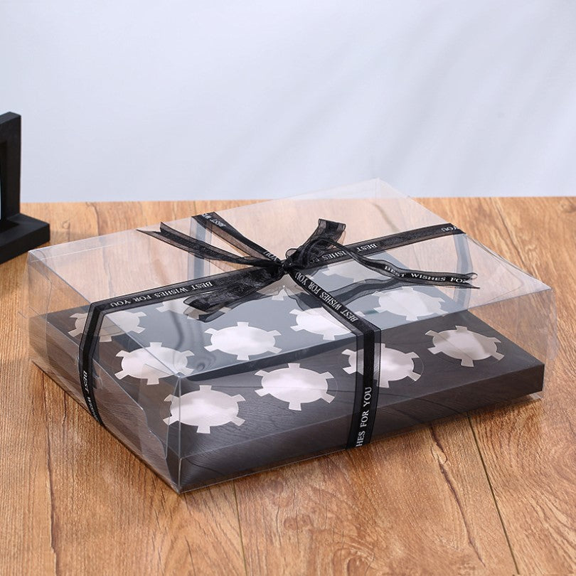 The image shows a clear cupcake box designed to hold 12 portions. The box features a transparent top with a decorative black ribbon tied around it, which has the inscription "Best Wishes for You." Inside, the base of the box is black and holds 12 circular cupcake holders. The box is resting on a wooden surface with a softly blurred background.