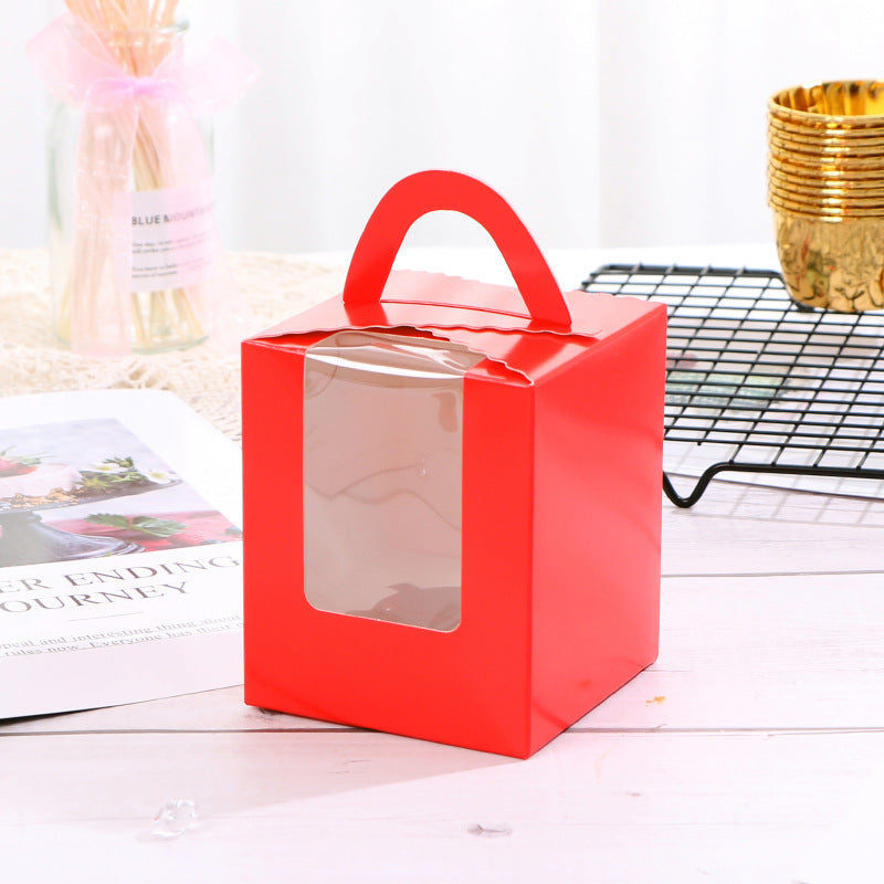 A bright red cupcake box designed for single portions. The box features a handle on top and a clear window on the front, allowing visibility of the contents inside. It is placed on a light wooden surface alongside a magazine and a wire rack in the background.