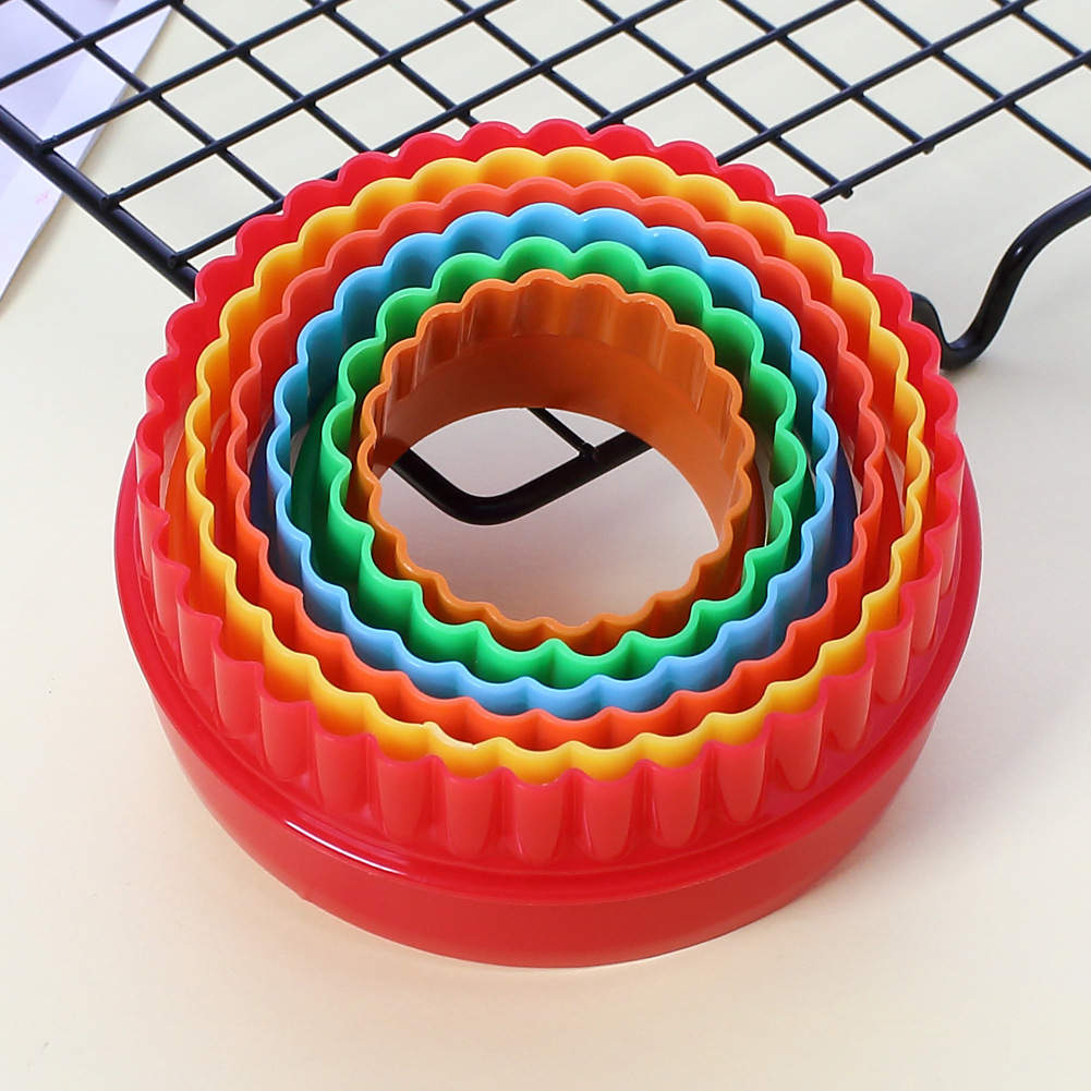 A round cookie cutter set consisting of six nested cutters in various colors. The outer cutter is red, followed by yellow, orange, green, light blue, and dark blue. The cutters are sitting on a light surface with a black wire cooling rack in the background.