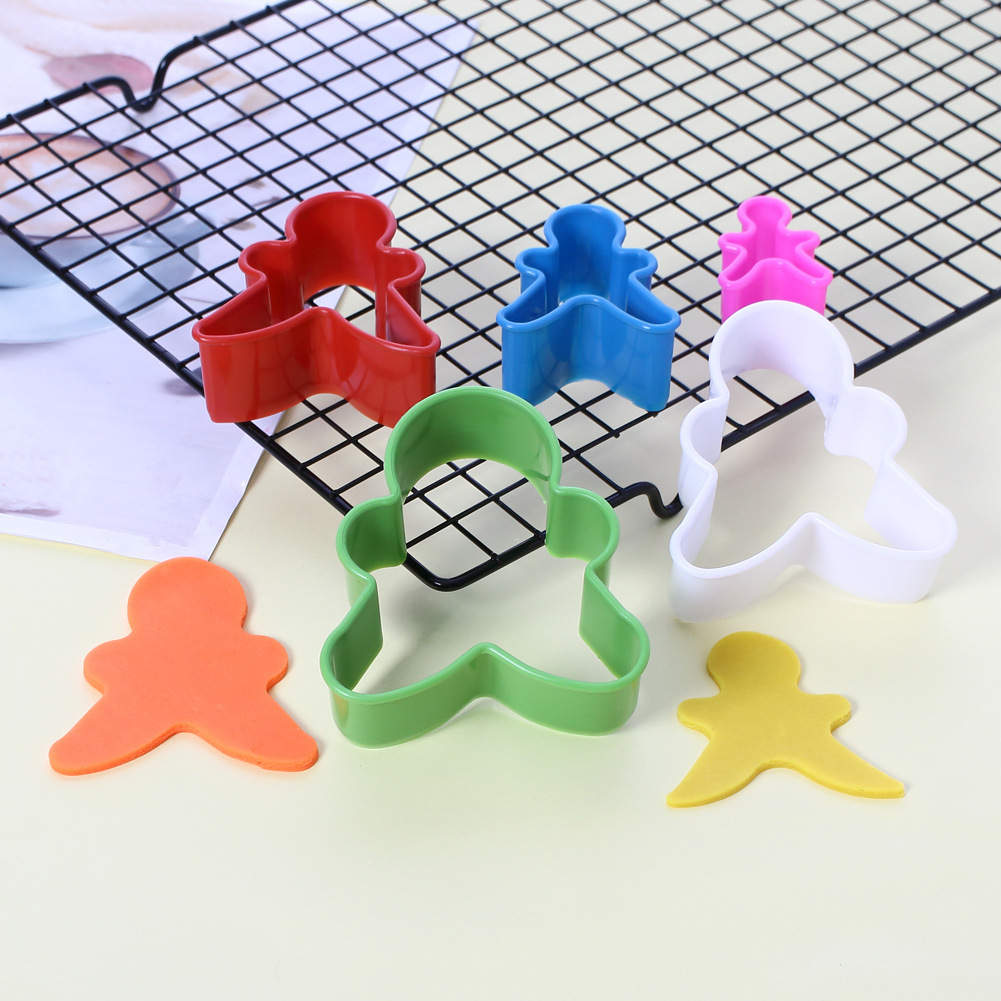 A set of five gingerbread man cookie cutters in various colors is displayed on a cooling rack. The cutters include red, blue, pink, green, and white. There are also two shaped cutouts in orange and yellow on a pale yellow surface next to the cutters. The arrangement showcases the playful design of the cookie cutters, ideal for making gingerbread man cookies.