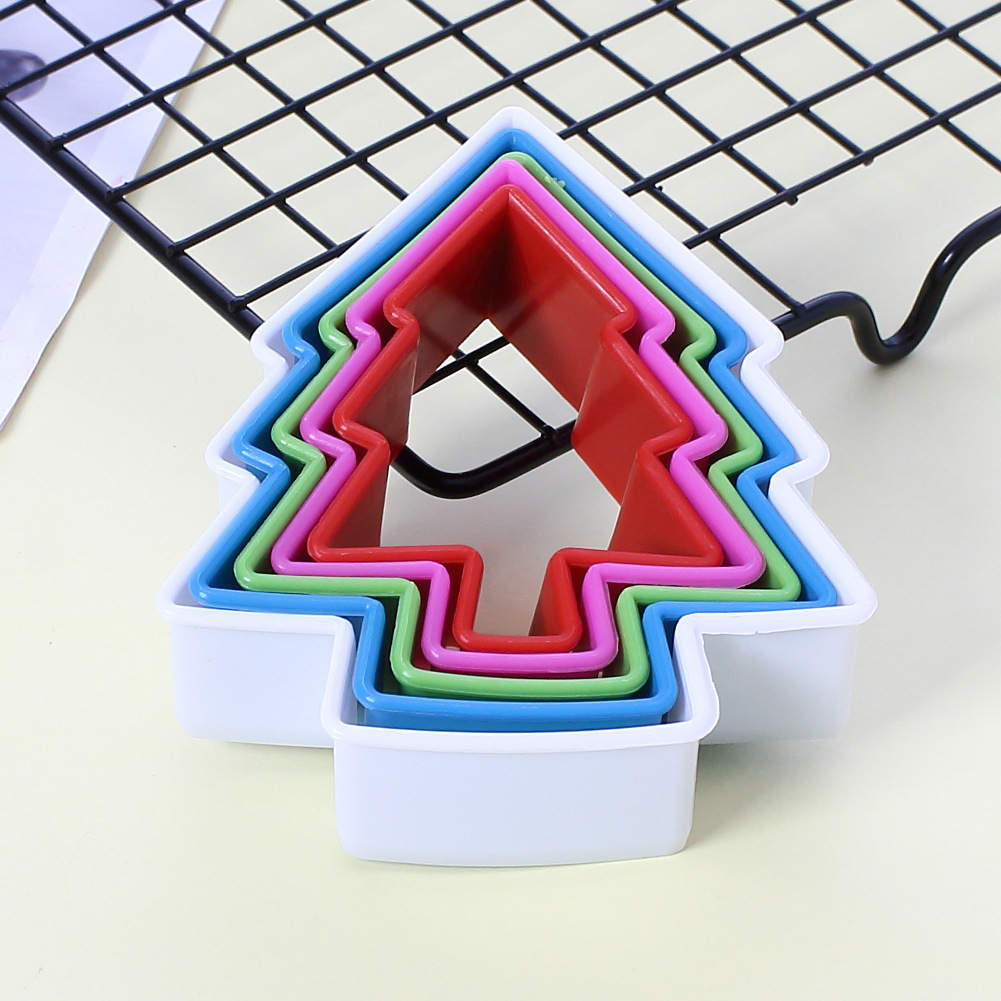 A set of five cookie cutters shaped like trees, stacked together. The cookie cutters are made of colorful plastic, with the outer layer being white, followed by green, pink, red, and blue layers in a nested arrangement. They are placed on a light-colored surface, with a wire cooling rack partially visible in the background.