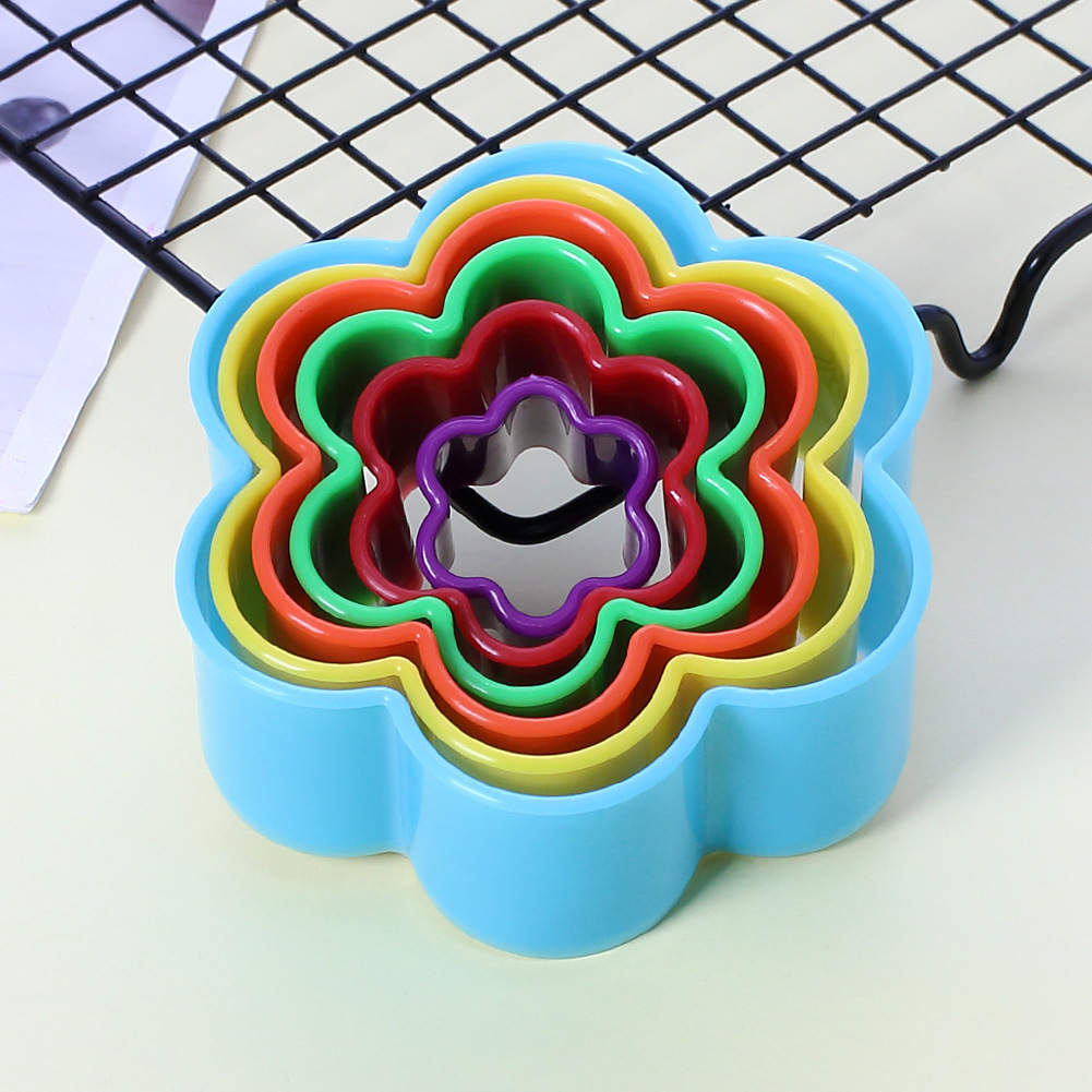 A flower cookie cutter set consisting of six colorful plastic cutters, stacked inside each other. The outermost cutter is light blue, followed by yellow, red, green, purple, and a small black cutter at the center. The background features a wire cooling rack and a light-colored surface.