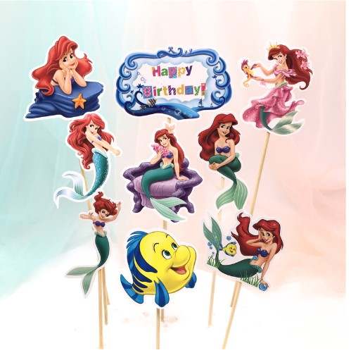 A group of cake toppers featuring various images of a mermaid character with red hair, styled in different poses. One topper displays the text "Happy Birthday" within a decorative border. Additional toppers include the mermaid sitting on a starfish, swimming, and in a pink dress, along with a yellow fish character. All toppers are mounted on sticks for decoration.