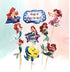 A group of cake toppers featuring various images of a mermaid character with red hair, styled in different poses. One topper displays the text "Happy Birthday" within a decorative border. Additional toppers include the mermaid sitting on a starfish, swimming, and in a pink dress, along with a yellow fish character. All toppers are mounted on sticks for decoration.