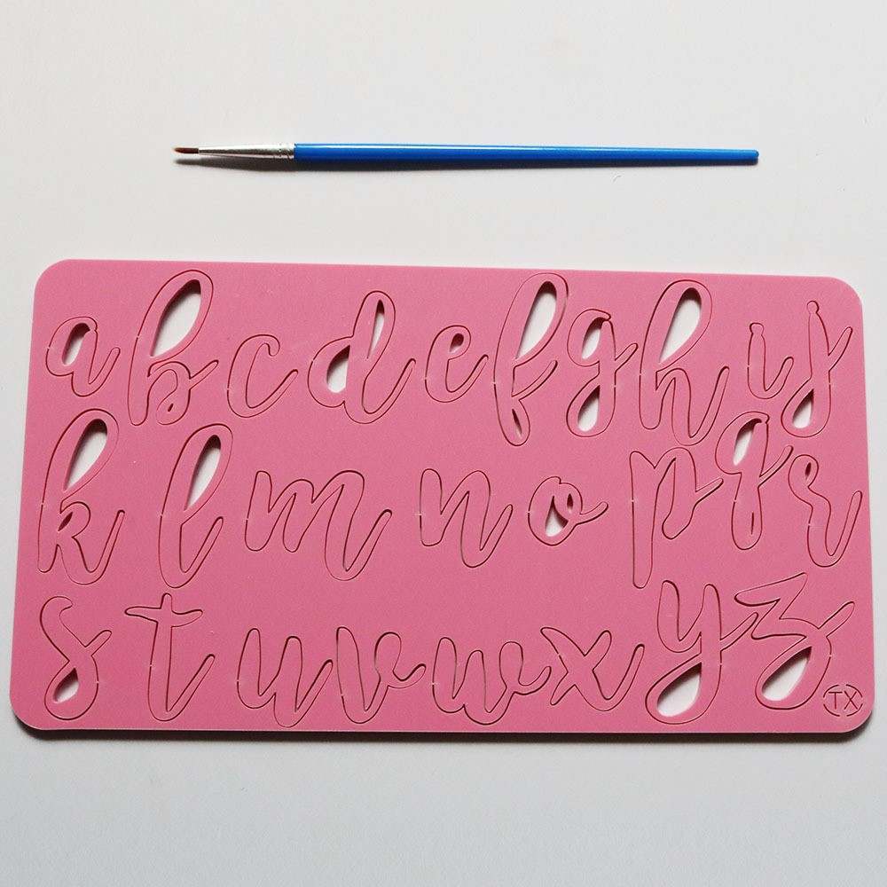 The image features a pink acrylic cake stamp with lowercase letters of the alphabet, designed in a cursive style. The letters are cut out of the stamp for easy use in cake decorating. A small blue paintbrush is positioned above the stamp, indicating that it may be used for filling the letters with icing or paint. The background is a light color, providing contrast to the pink stamp.