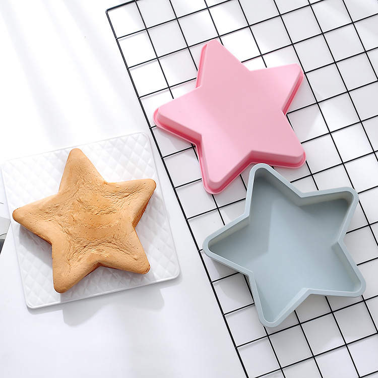 A flatlay image featuring three star-shaped silicone moulds: one pink and one light blue, placed above a brown star-shaped cake on a white textured surface. The background includes a black and white grid pattern.