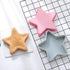A flatlay image featuring three star-shaped silicone moulds: one pink and one light blue, placed above a brown star-shaped cake on a white textured surface. The background includes a black and white grid pattern.