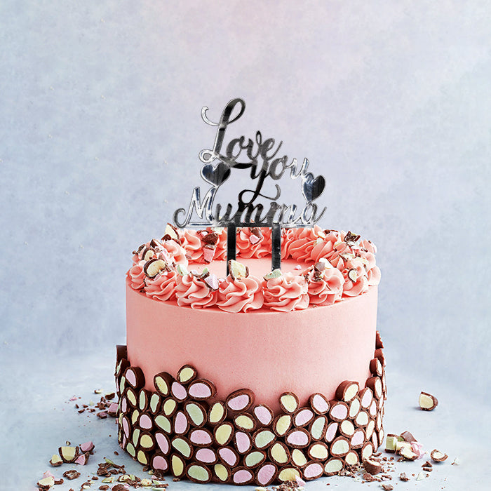 A pink frosted cake decorated with rosettes along the edge and a chocolate garnish at the base. At the top, a customized cake topper reads "Love you Mumma" in a stylish font, accompanied by small heart shapes. The background is light and neutral, enhancing the cake&
