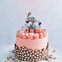 A pink frosted cake decorated with rosettes along the edge and a chocolate garnish at the base. At the top, a customized cake topper reads "Love you Mumma" in a stylish font, accompanied by small heart shapes. The background is light and neutral, enhancing the cake&
