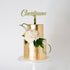 A two-tiered cake with a shiny gold finish, topped with a customized name topper reading "Christiaan." The cake is adorned with white flowers and green foliage, creating an elegant presentation on a white cake stand against a plain white background.