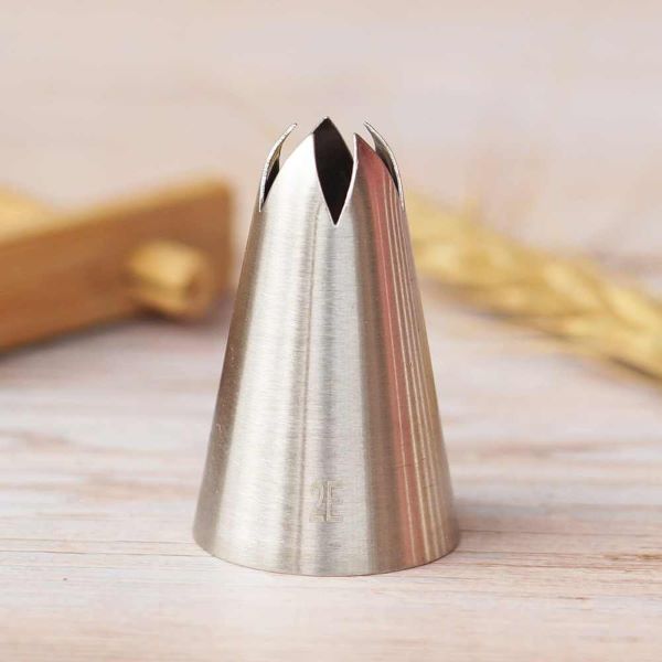 A stainless steel piping nozzle with a tapered, cone shape and multiple slits at the top, labeled "2E." The nozzle rests on a wooden surface, with a blurred background featuring a wooden utensil and some wheat stalks.