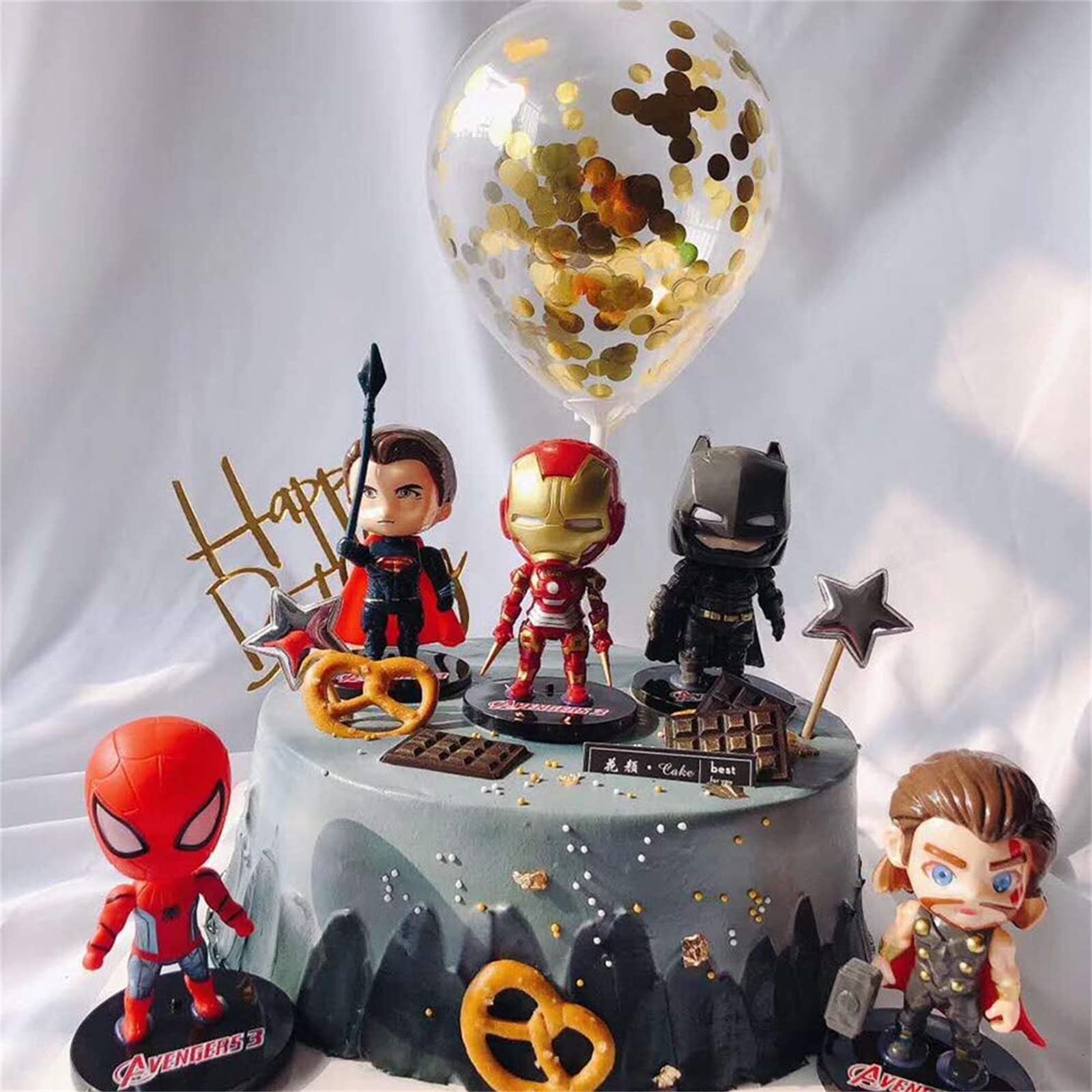 A decorated birthday cake featuring five colorful mini cartoon superhero figurines from the Avengers. The characters displayed include Iron Man, Spider-Man, a superhero with a spear, Batman, and a figure resembling Thor. The cake is gray with a textured surface and adorned with chocolate pieces, pretzels, and gold confetti. Above the cake, there is a clear balloon filled with gold confetti and a "Happy Birthday" sign made of gold lettering. The setting has a soft, light background.