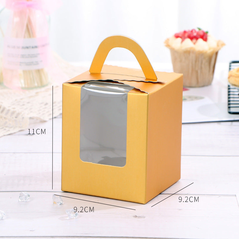A yellow cardboard cupcake box designed for a single portion, featuring a transparent window on the front. The box measures 11 cm in height and 9.2 cm in width and depth. In the background, there is a blurred image of a cupcake and some decorative elements.