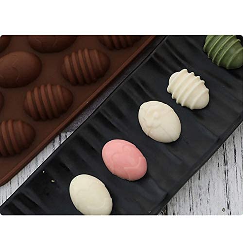 A silicone mould designed for making egg-shaped treats, featuring 15 cavities in a dark brown color. Next to the mould, there is a black serving plate displaying five colorful egg-shaped chocolates: two brown, one pale pink, one white, and one green, each with a ribbed texture. The background shows a light-colored wooden surface.