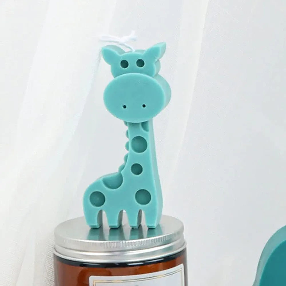 A teal silicone mould shaped like a giraffe, featuring large round spots and a friendly expression, stands atop a silver-lidded jar. The giraffe has a long neck and distinct features, with its ears and small horns visible at the top. The background is light and airy, suggesting a bright setting.