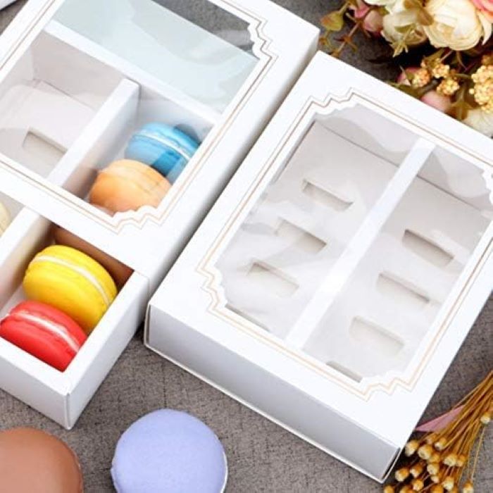 The image features a product called "window macaron box 10 portions." There are two white boxes displayed, each with a clear window. One box contains four brightly colored macarons: red, yellow, orange, and blue. The other box is empty, showing six compartments designed for macarons. Soft pastel-colored macarons, like a lavender one, are also visible on the surface beside the boxes. The background includes decorative dried flowers, enhancing the overall presentation.