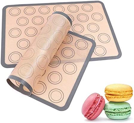 A rolled-up silicone baking mat is partially unrolled over a flat baking surface, featuring circular guides for placing items like macarons. Next to the mat, there are three colorful macarons in pink, yellow, and green.