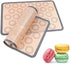 A rolled-up silicone baking mat is partially unrolled over a flat baking surface, featuring circular guides for placing items like macarons. Next to the mat, there are three colorful macarons in pink, yellow, and green.