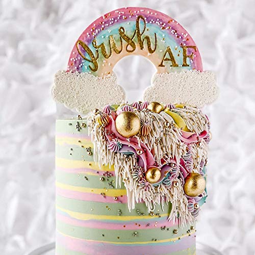 The image features a beautifully decorated cake topped with a colorful rainbow and fluffy clouds. The rainbow has the words "Brush AF" written in gold lettering, and it sits above a cascading design of icing. The cake itself is decorated with pastel stripes, and the icing drapes down in a textured, artistic manner, adorned with various decorative elements including golden spheres and colorful swirls. The background is a soft, neutral fabric that adds to the overall aesthetic.