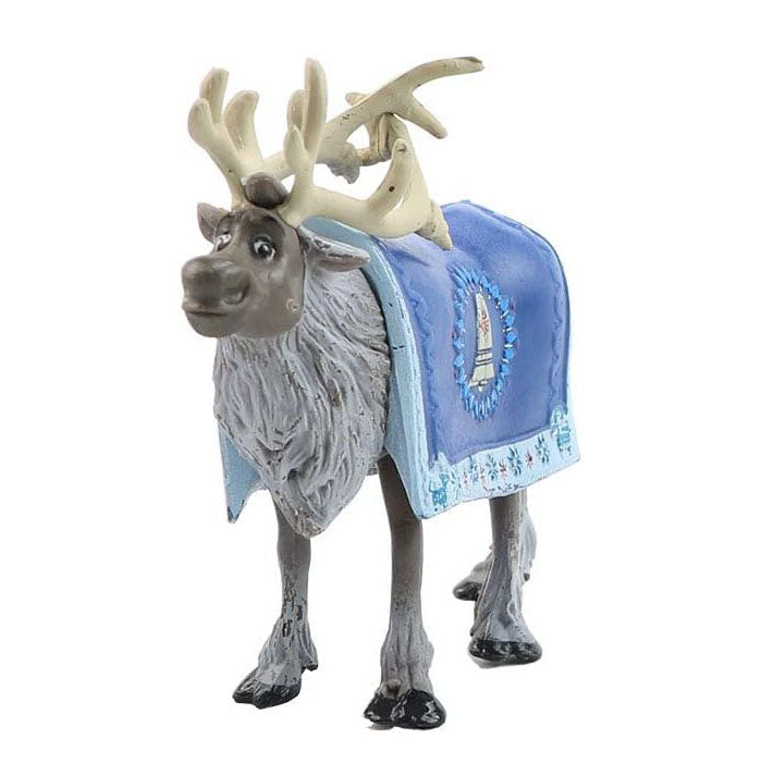 A toy figure of a reindeer with large antlers and a detailed fur texture. It has a blue blanket draped over its back, featuring intricate patterns. The figure is standing on a flat surface, showcasing its playful and friendly expression.