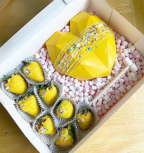 An image shows a heart-shaped piñata mold in bright yellow, embellished with colorful sprinkles and a white candy stick. Surrounding the piñata are several chocolate-dipped strawberries, also yellow with decorative white drizzle and small, colorful sprinkles, placed in paper cups. The entire arrangement is set in a box filled with pink and white marshmallow-like candies.