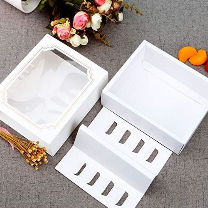 A white macaron box designed to hold 10 portions, featuring a clear window on the lid. The box is partially open, showing a compartment insert with cutouts for holding macarons securely. Next to the box, there are small flowers, adding a decorative touch to the scene. The background is a textured gray surface.