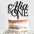 A close-up view of a layered, semi-naked cake topped with a decorative cake topper. The topper features the text "Alia is ONE" in an elegant, modern font. The cake has a rustic appearance with visible layers of cake and frosting, surrounded by small white flowers on the surface. The background is a light gray, enhancing the focus on the cake and topper.
