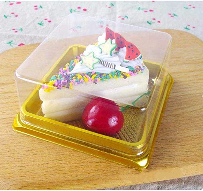 A small dessert displayed in a clear plastic box. The dessert resembles a slice of cake, topped with colorful sprinkles, white icing, and small decorative elements including green stars and red shapes that appear to be strawberries. Below the cake slice, there is a shiny red cherry placed on a textured golden base. The box is set on a wooden surface with a light floral fabric underneath.