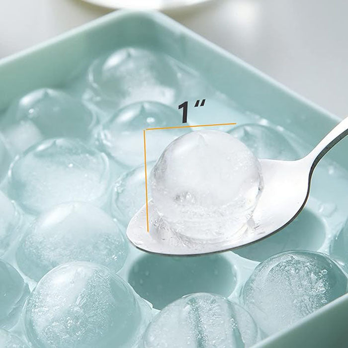 A round ice cube tray filled with spherical ice cubes. One ice cube is being lifted with a spoon, showing its size of approximately 1 inch. The tray is light blue, and the ice cubes are clear and slightly translucent.