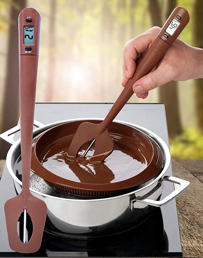 An image features a digital spatula candy thermometer, which is being used to stir a pot of melted chocolate. The thermometer has a digital display showing a temperature reading. The spatula is designed for cooking and features a flexible head for mixing. The background shows a natural setting, suggesting an outdoor atmosphere, while the foreground includes a stainless steel pot placed on a stovetop.
