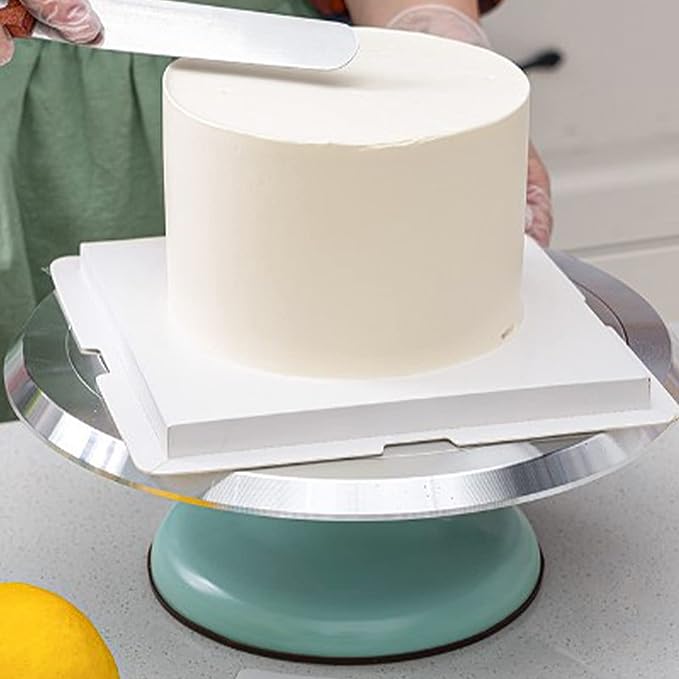 An aluminium revolving turntable is shown in the image, featuring a smooth surface. A round, frosted cake is placed on a white cake board atop the turntable. A person, wearing gloves, is using a spatula to smooth the icing on the top of the cake. The turntable has a mint green base, and the setup is on a light-colored countertop.