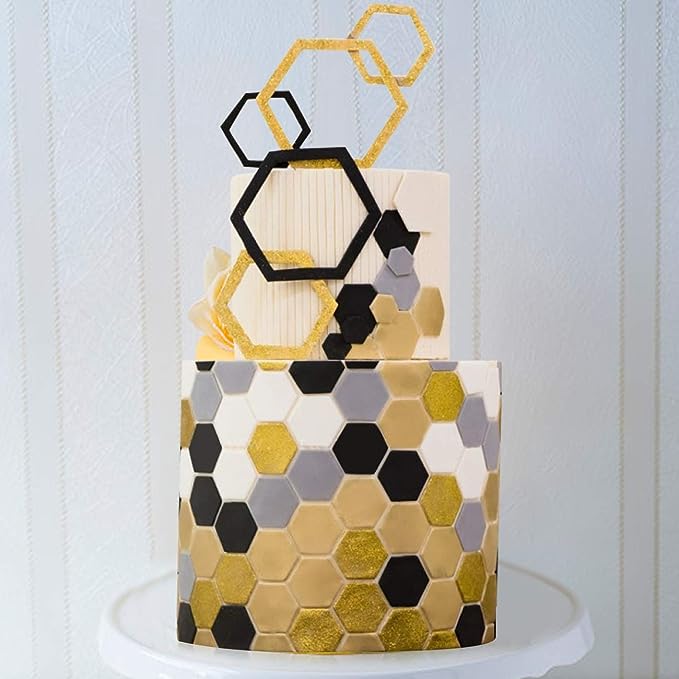 A decorative cake featuring hexagon patterns in shades of black, gold, and gray. The cake is adorned with hexagon-shaped toppers in gold glitter and a black hexagon design. The background is a subtle striped pattern, and the cake is placed on a white pedestal.