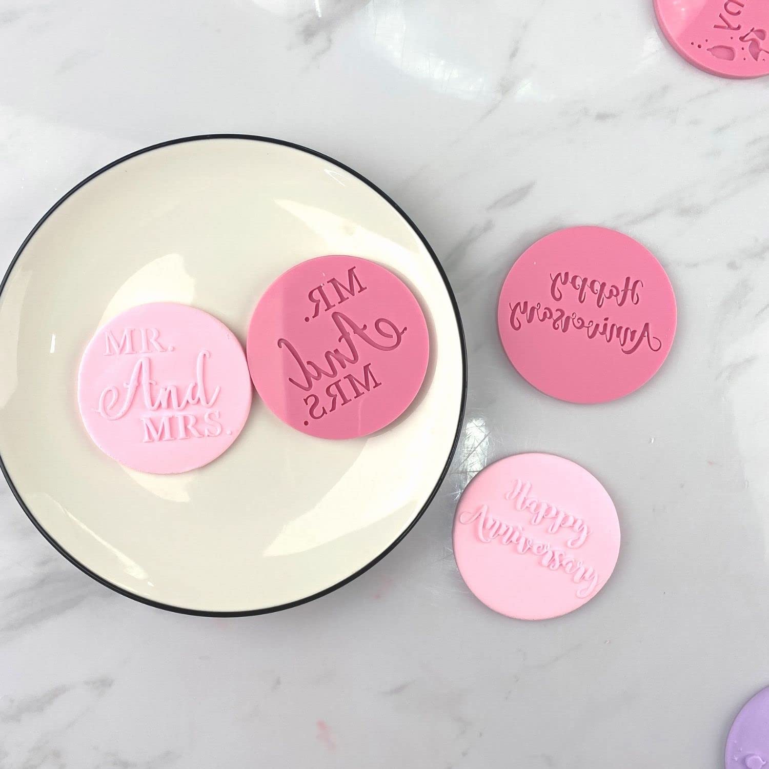 A collection of circular fondant embosser cookie stamps is displayed on a white plate. The stamps feature various phrases: one is pink with "MR. & MRS." embossed, another is pink with "And" in a decorative font, and a third is pink with "Happy Anniversary" text. There are also two additional pink stamps with similar designs, but their phrases are obscured. The background is a light marble surface.