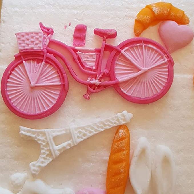 The image shows a silicone mould featuring a pink bicycle with a basket, positioned on a light-colored surface. Surrounding the bicycle are several themed elements, including a white Eiffel Tower, an orange croissant, a pink heart, an orange baguette, and a white slipper or flip-flop. The mould captures various Parisian motifs in a playful and colorful design.