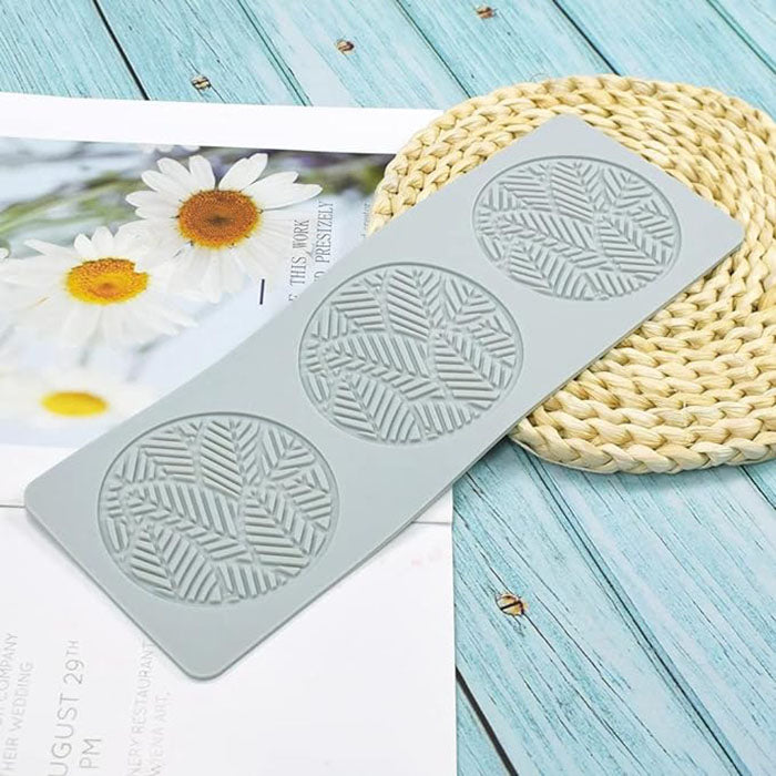A gray silicone mold designed for crafting, featuring three circular cavities with an embossed leaf pattern. The mold is placed on a light blue wooden surface, alongside a woven placemat and a magazine showing a photo of daisies.