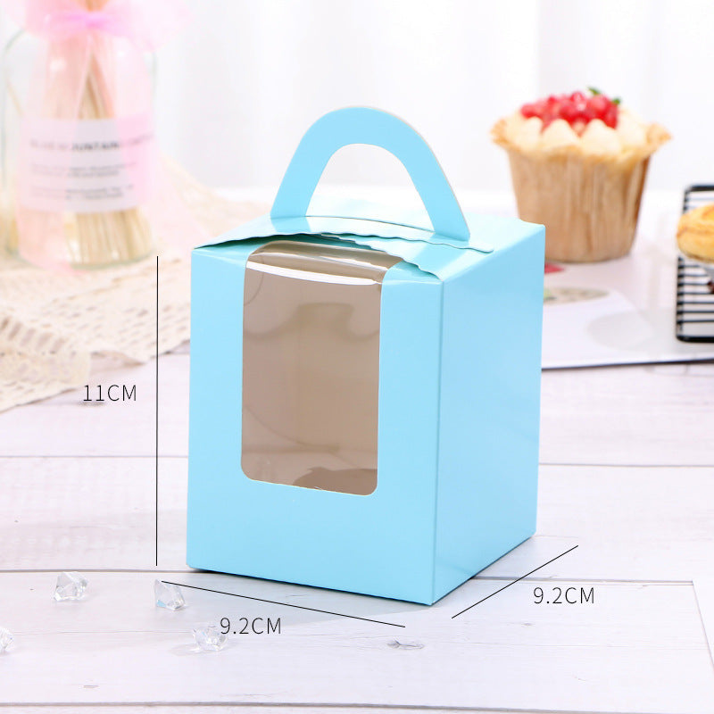 A light blue cupcake box with a handle sits on a white, wooden surface. The box has a clear window on one side, allowing visibility of the contents inside. The dimensions of the box are indicated: height is 11 cm and the width and depth are both 9.2 cm. In the background, there is a blurry image of a cupcake topped with red berries and cream, as well as a jar with decorative sticks, suggesting a setting for baked goods or a bakery display. Small crystal-like decorations are scattered around the box.